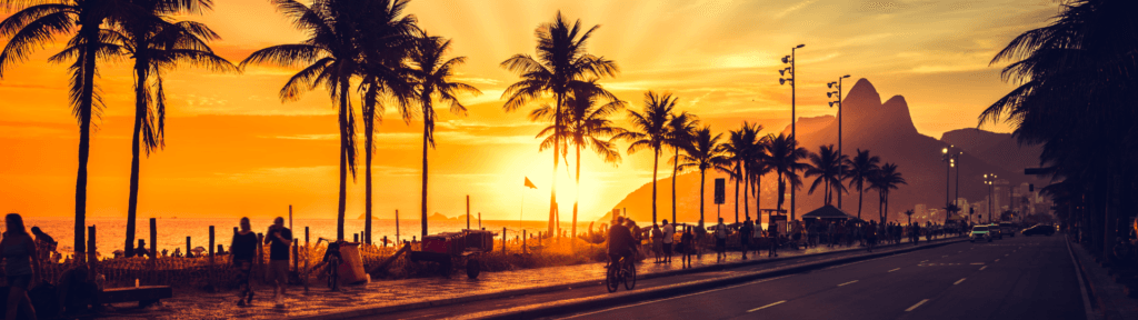 Localização estratégica no Rio transforma imóvel em resultado consistente Pôr do sol na orla do Rio de Janeiro com palmeiras, pessoas caminhando e o céu alaranjado, simbolizando localização como estratégia de valor no aluguel por temporada.